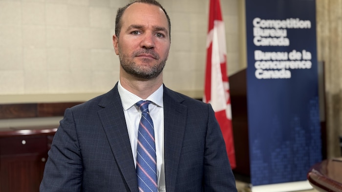 Anthony Durocher avec un air sérieux est face à la caméra. Le drapeau du Canada est en arrière-plan.
