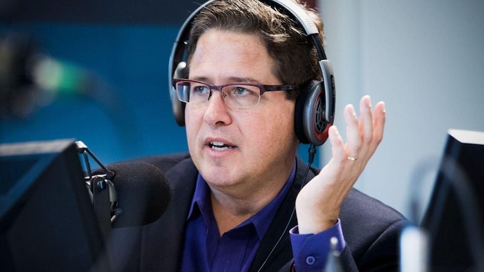 Un animateur de radio assis avec un casque d'écoute sur la tête, devant un micro. Il a la main gauche levée et parle.