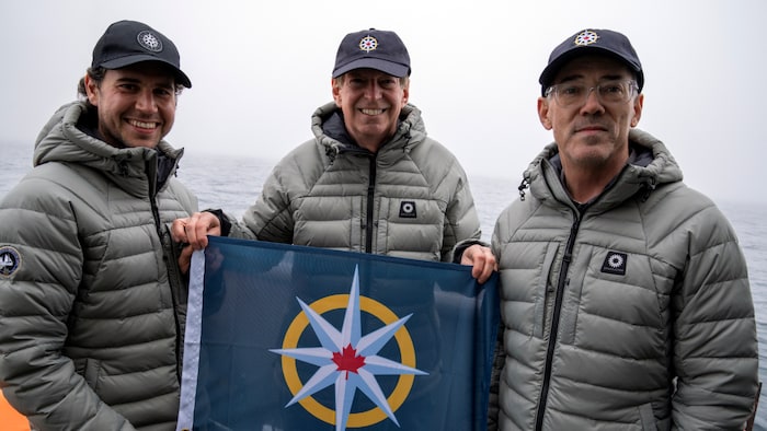 Antoine Normandin, John Geiger et David Mearns souriants sur un bateau, un drapeau naval à la main.