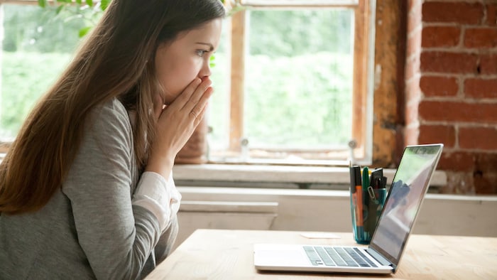 Une jeune femme semble inquiète et met les mains devant sa bouche en regardant l'écran de son ordinateur.