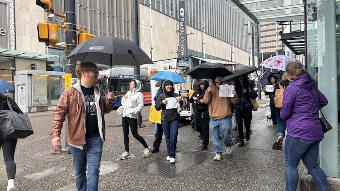 Un groupe de jeunes sous des parapluies marche sur un trottoir mouillé du centre-ville en tenant des affiches sur lesquelles est écrit « VOTE ».