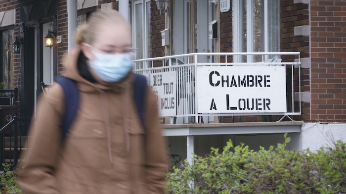 فتاة تضع قناع وجه واقياً تمرّ أمام مبنى سكني عليه لافتة تقول إنّ فيه غرفة للإيجار.
