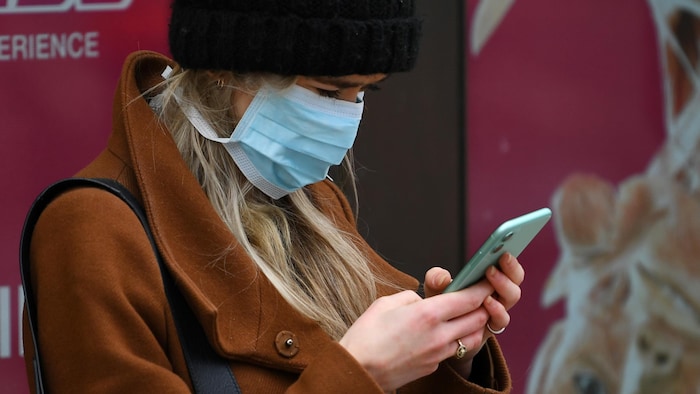 Une femme, coiffée d'une tuque en laine et le visage recouvert d'un masque médical, est penchée sur son téléphone cellulaire.