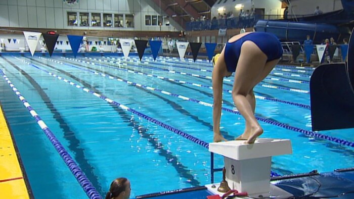 Une femme en maillot bleu est penchée sur le bloc de départ avant de plonger dans une piscine.