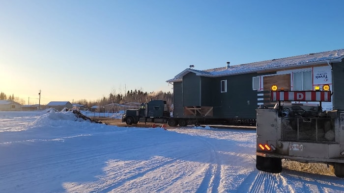 Une maison préfabriquée est transportée sur une remorque dans une communauté enneigée. Un camion l'accompagne.