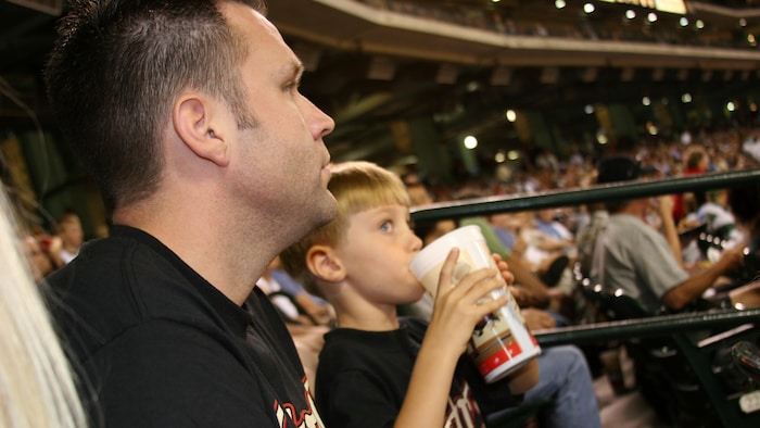 Un père et son fils dans les gradins d'un aréna, l'enfant boit une boisson sucrée