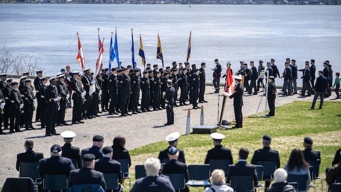 Des membres de la Marine royale canadienne en uniforme sont rassemblés sur le bord du fleuve à Trois-Rivières avec des drapeaux à la main. 