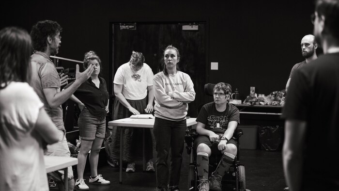 Sept personnes échangent en cercle dans un studio de théâtre dont une utilisant un fauteuil roulant électrique.