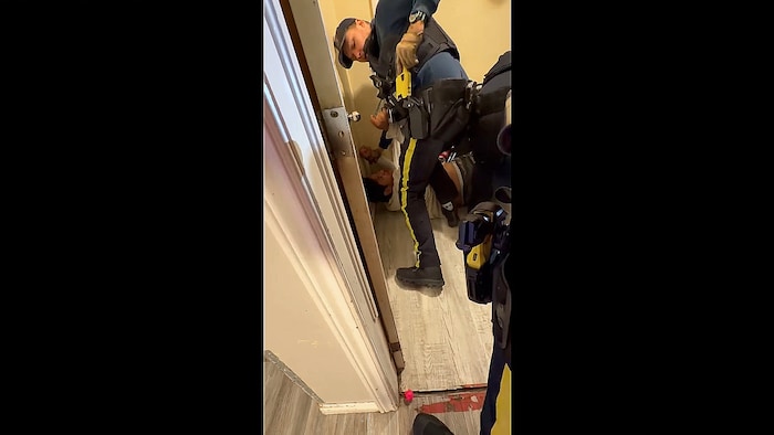 Un policier debout remettant un pistolet Taser jaune dans l'étui à sa ceinture, pendant qu'un homme en civil est couché sur le dos, sur le plancher.