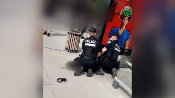 Deux policiers agenouillés, dont un pose son genou sur le cou de la personne arrêtée, près d'un abribus.