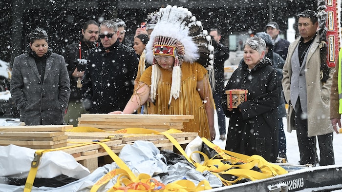 Une cheffe autochtone se penche devant des caisses de bois contenant les artefacts  sous la neige. 