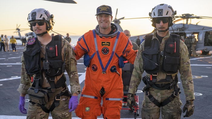 L'astronaute Jeremy Hansen reçoit de l'aide pour quitter le pont de la part de deux personnes en uniforme.