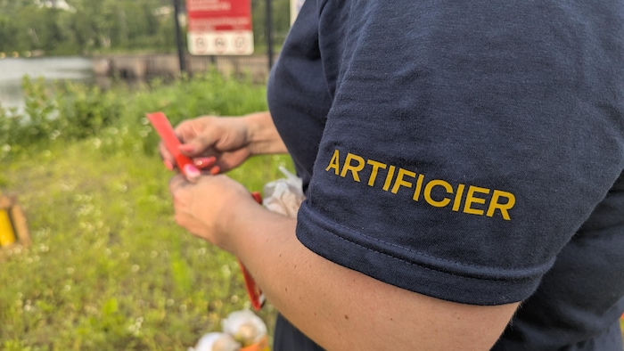 Gros plan sur la manche du chandail de Jennifer, où il est inscrit « artificier ». Elle place un plastique sur une mèche.