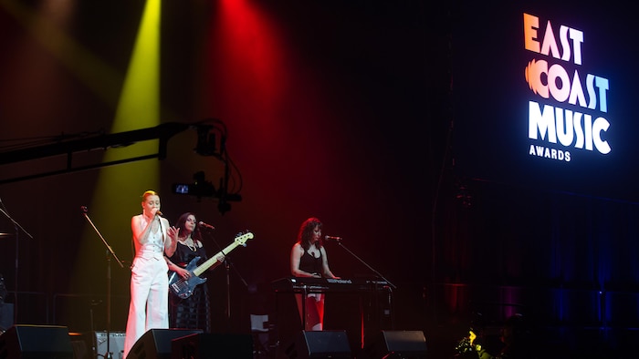 Une jeune femme chante sur la scène, accompagnée de deux autres artistes féminines à la guitare et au clavier.