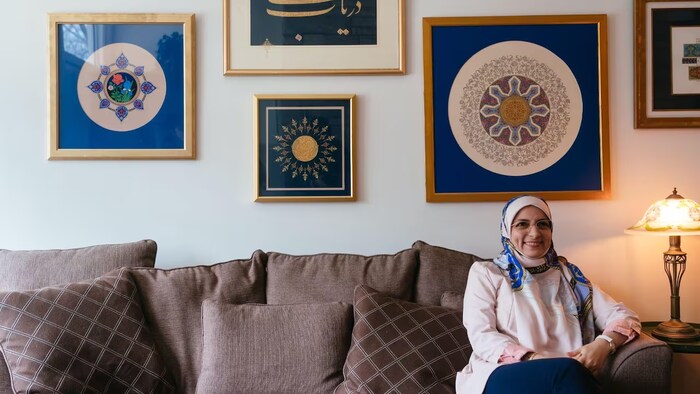 المُذهِّبة الكندية الإيرانية مريم نديمي جالسة على كنبة في منزلها في مونتريال، ورسوماتها معروضة على الجدار خلفها.
