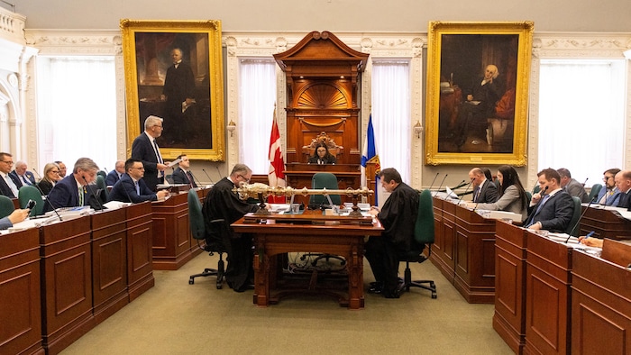 Les députés sont assis des deux côtés de l'allée centrale de la législature. La présidente est assise au centre. Le ministre des Finances, à gauche, est debout.