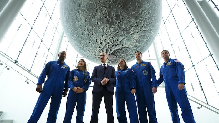 Le premier ministre canadien et les astronautes se tiennent debout, derrière eux se situe une maquette de la Lune. 