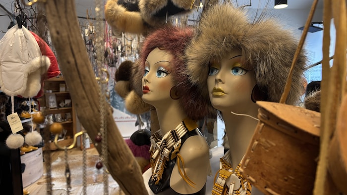 Deux bustes de mannequins féminins sont présentés côte à côte en gros plan. Ils portent une coiffe volumineuse en fourrure ainsi qu'un collier plastron composé de perles allongées et de lanières de cuir. 