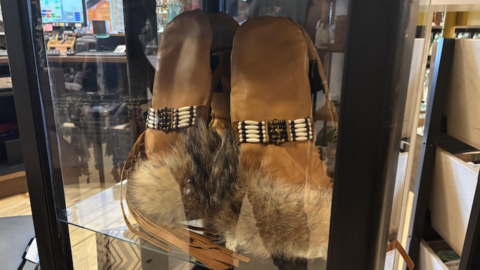 Vue rapprochée, à travers une vitrine en verre, d'une paire de mitaines hautes en cuir ornées de bordures en fourrure dense au niveau des poignets.