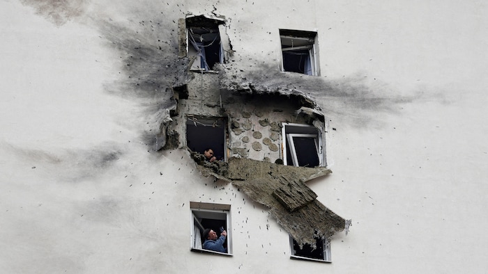 Un immeuble résidentiel endommagé avec, à ses fenêtres, des personnes qui prennent des photos avec leur téléphone cellulaire. 