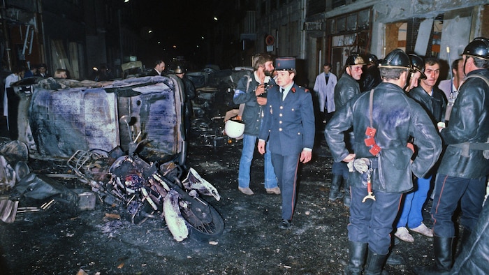 Des pompiers sont debout près d'une voiture et d'une moto incendiées dans une rue.