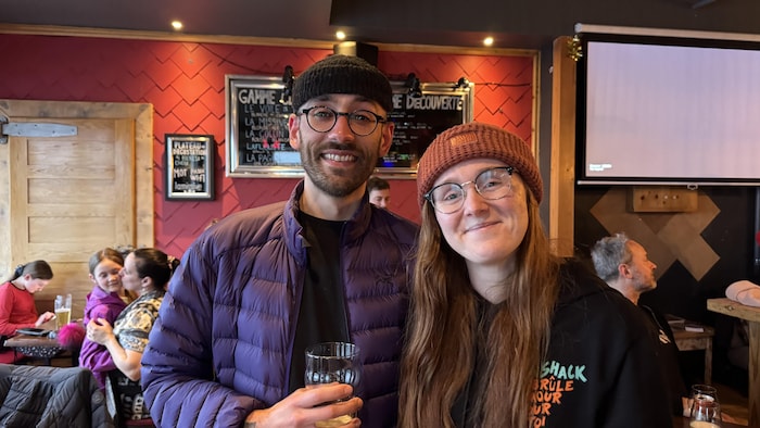 Un couple avec une tuque et des lunettes regarde l'objectif en souriant, un verre à la main.