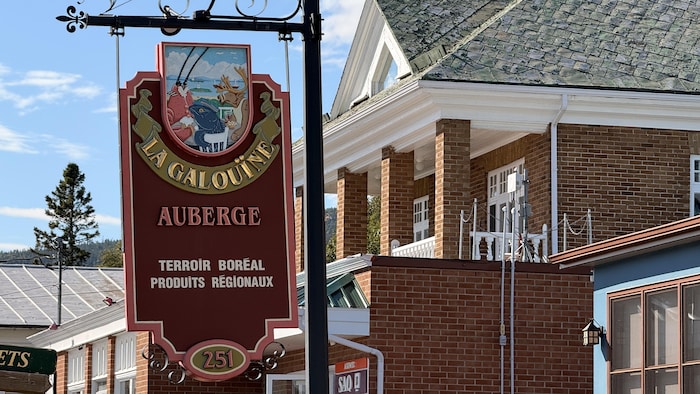 Un gros plan d'une enseigne commerciale ornée et suspendue en fer forgé. Le panneau présente le texte « LA GALOUÏNE AUBERGE » et un logo illustré représentant des paysages et de la faune.