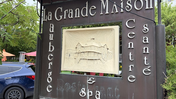 La Grande Maison, auberge & Spa, Centre Santé. La trace du mot restaurant apparaît toujours, même si les lettres qui le formaient ont été retirées. Au centre de l'affiche est installée une sculpture sur bois, représentant la maison.
