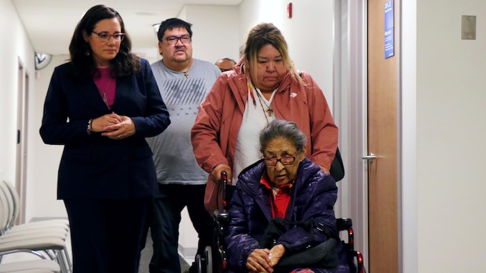 Trois personnes marchent en accompagnant une femme âgée en fauteuil roulant.