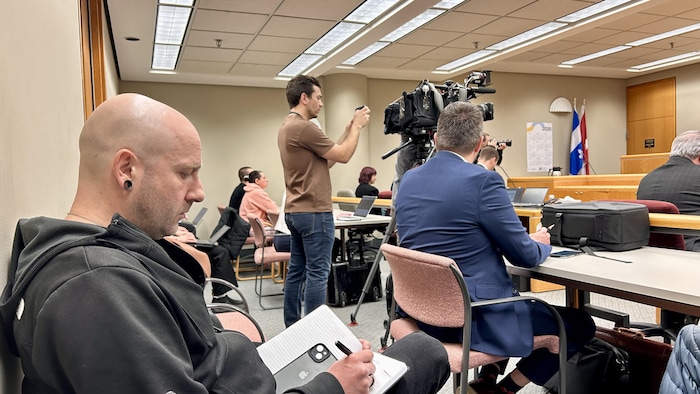 Dans une salle d'audiences où se trouve plusieurs personnes et des représentants des médias, un homme prend des notes, assis à l'arrière de la salle. 