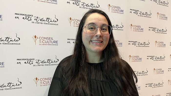 Audrey Desrochers pose devant une affiche des Prix d'excellence en arts et culture, souriante.