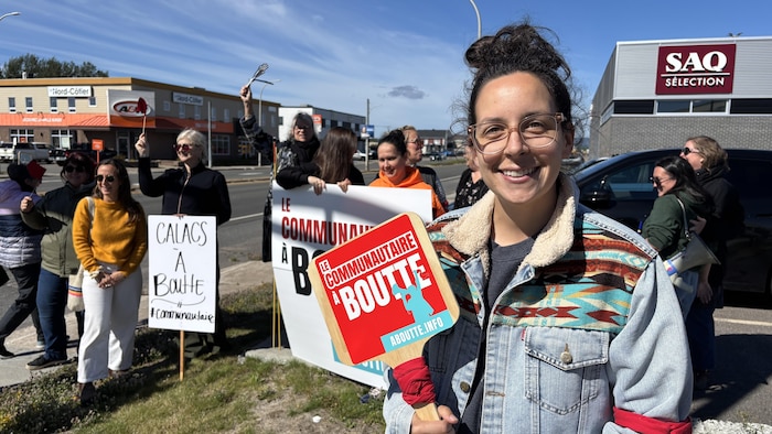 Portrait en plan moyen d'une femme souriante portant des lunettes de vue et une veste en jean à motifs. Elle tient devant elle un panneau rigide muni d'une poignée sur lequel est inscrit « LE COMMUNAUTAIRE À BOUTTE ».
