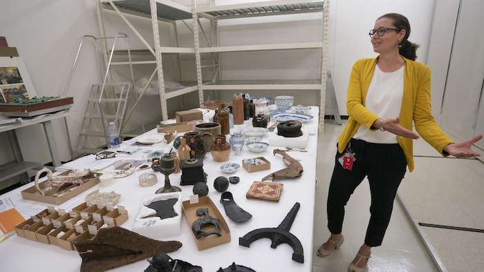 Une femme se tient devant une table contenant des artéfacts de différentes époques trouvés dans le sous-sol montréalais.