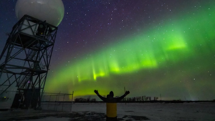 Une personne de dos se tient les bras tendus vers le ciel en face d'une aurore boréale.