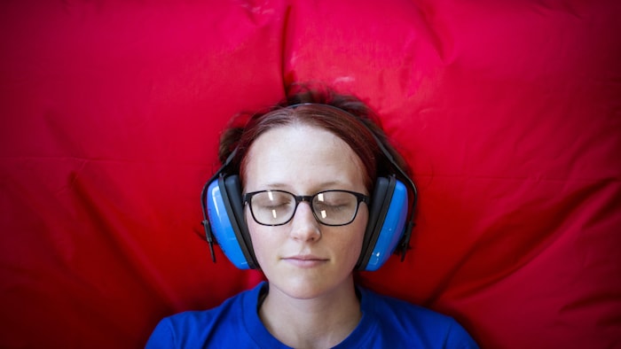 Portrait rapproché de Kayla Tellier, une jeune fille aux yeux fermés allongée sur un oreiller rouge avec un casque de réduction sonore sur les oreilles

