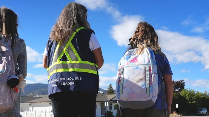 Deux enfants portant un sac à dos se tiennent à côté d'une femme vêtue d'un dossard, de dos, à un coin de rue.