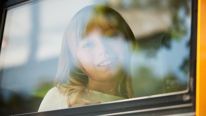 Le visage d'une petite fille assise dans un autobus scolaire apparaît à la fenêtre du véhicule. Elle est photographiée de l'extérieur.