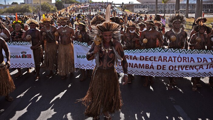 Des Autochtones brésiliens en costumes traditionnels marchent en tenant une banderole.