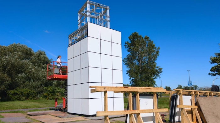Deux ouvriers sur une nacelle élévatrice assemblent une grande structure métallique dans un parc au bord de l'eau.
