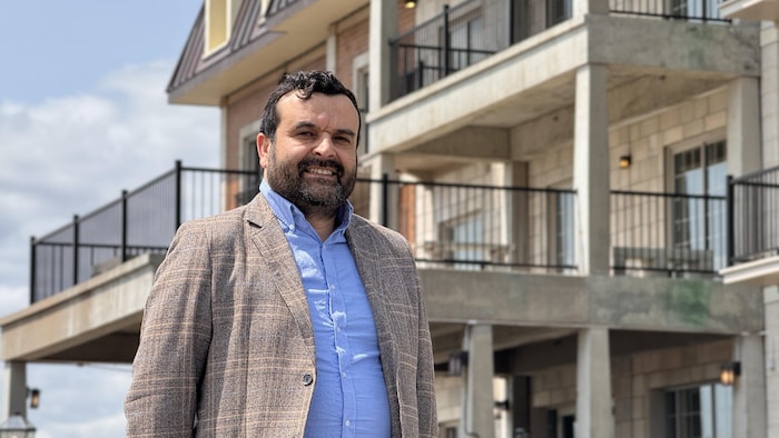Un homme en complet sourit à la caméra devant un bâtiment.