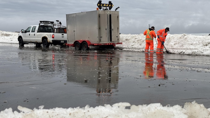 Deux travailleurs en tenue de sécurité orange déglacent un terrain inondé près d'une remorque de signalisation.