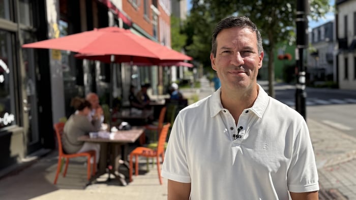 Un homme souriant en t-shirt blanc est debout devant la terrasse extérieure d’un café sur un trottoir.