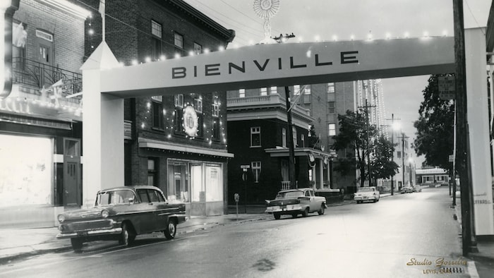 Un portique illuminé arborant le mot « BIENVILLE » surplombe une rue bordée de bâtiments et de voitures stationnées.