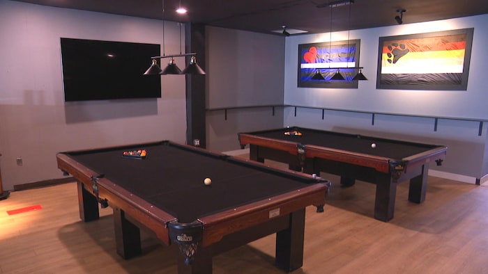 Deux tables de billard. Un drapeau signifiant la communauté cuir et un drapeau bear sur un mur. Un écran de télévision éteint sur un autre mur.