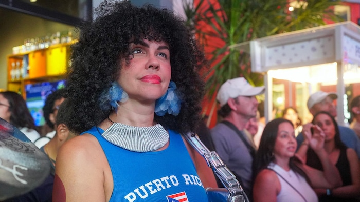 Des gens regardent un écran hors champ, souriant. Une femme au premier rang porte un chandail avec le drapeau portoricain.
