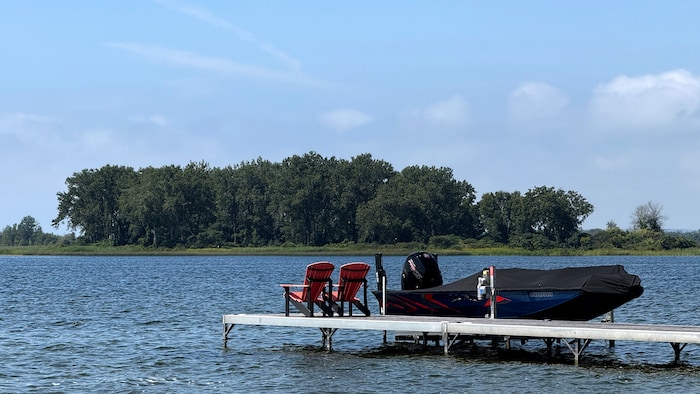 Un quai avec deux chaises rouges de type Muskoka, un bateau sur l'eau de la baie, l’autre côté de l’eau, on peut apercevoir l'île Bald.
