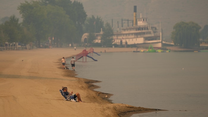 Des personnes sont assises sur la plage tandis que la fumée de l'incendie de Bald Range enveloppe le lac Okanagan, à Penticton, en Colombie-Britannique, le dimanche 9 août 2026. 