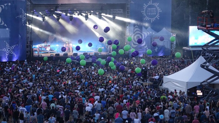 Des ballons naviguent au-dessus d'une foule de personnes qui assistent à un spectacle extérieur. On voit une grande scène avec des projecteurs de lumière.