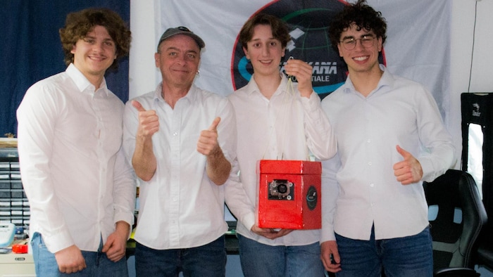 Trois étudiants et M. Nadeau posent avec une petite boîte rouge, la sonde, devant une toile qui fait la promotion de Nanikana. Les trois arborent un grand sourire.