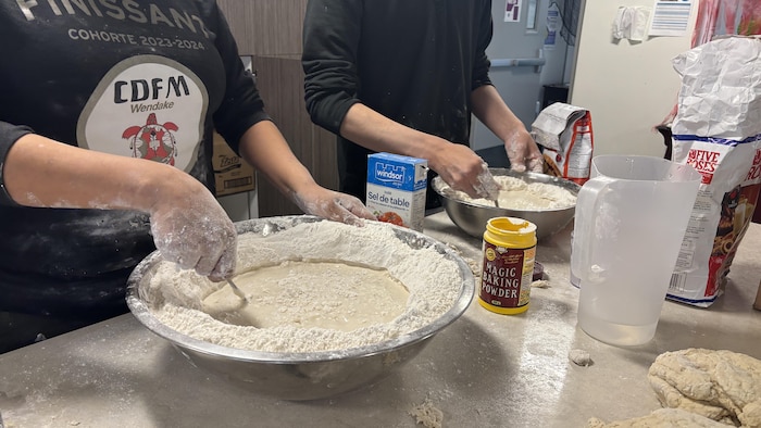 Deux personnes sont debout au-dessus d'un comptoir couvert de farine. Au premier plan, des mains mélangent de l'eau et de la farine dans un grand cul-de-poule métallique. À côté, on aperçoit une boîte de poudre à pâte, un contenant de sel et un grand sac de farine. En arrière-plan, une deuxième personne prépare un autre bol de pâte.